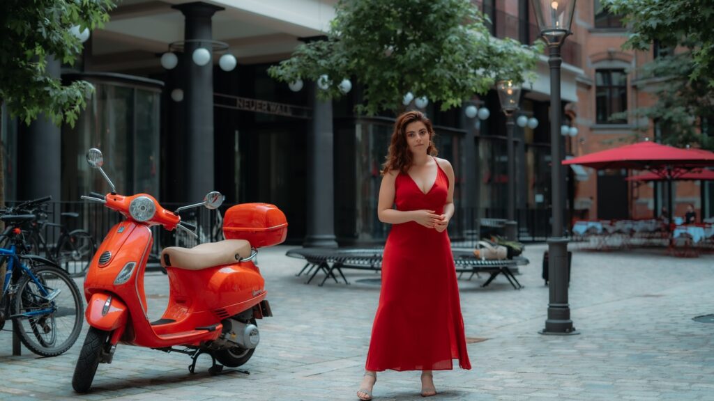 woman in red sleeveless dress standing beside red motor scooter during daytime